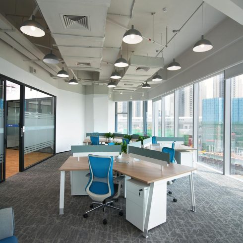 Bright modern office with floor-to-ceiling windows, clustered wooden desks, blue chairs, and exposed ceiling ducts.