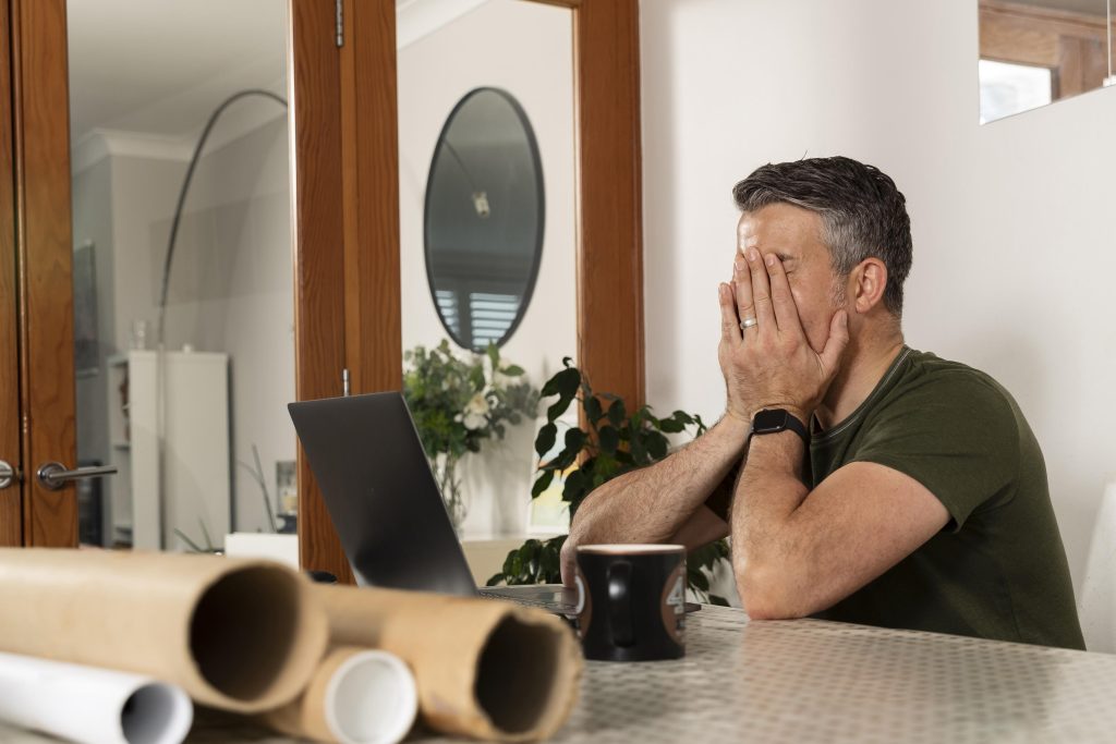 Stressed professional at a desk with blueprints and a laptop, illustrating the frustration of hidden engineering costs.